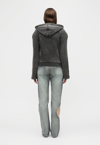 Άτομο με σκούρο γκρι μπουφάν με κουκούλα και ριμπ λεπτομέρειες στους ώμους, και ανοιχτό γκρι τζιν με κυκλική εγκοπή στο δεξί πόδι, στέκεται γυρισμένο προς τα πίσω.