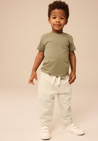 Zeleno kratko majico s kratkimi rokavi in žepom, kombinirano z belimi hlačami s črtami ter svetlimi supergami z ježkom. Nevtralna ozadje.