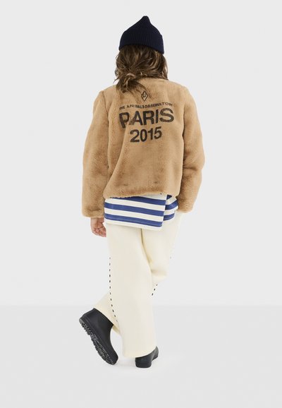 Tan faux fur-jakke med sort trykt tekst "PARIS 2015." Laget med en hvid og blå stribet skjorte og cremede bukser med stikninger. Sorte støvler.