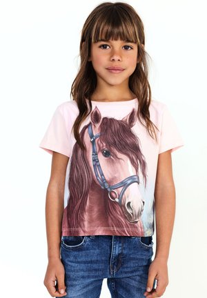 Ragazza con lunghi capelli castani indossa una maglietta rosa chiaro con una grande testa di cavallo marrone con capezza blu, abbinata a jeans blu.