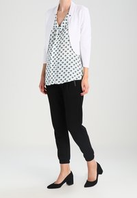 White cropped jacket over a green-and-white patterned blouse, paired with black tapered pants and black block-heeled shoes.