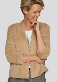 Beiger, flauschiger Cardigan mit Schalkragen und dreiviertel Ärmeln. Weiche Textur und lockere Passform, kombiniert mit einem grafischen T-Shirt darunter.