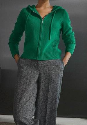 Vihreä hupullinen vetoketjuhuppari, jossa resorireunukset hihoissa ja helmassa, yhdistetty harmaisiin, kuvioituihin, leveälahkeisiin housuihin. Taustalla hillitty miljöö.