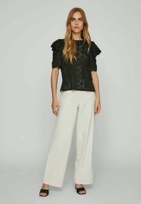 Γυναίκα με μαύρη δαντελωτή μπλούζα κοντομάνικη με ριχτές ώμους, λευκά παντελόνια φάρδυ, και μαύρα ανοιχτόκανα παπούτσια, που στέκεται μπροστά σε απλό φόντο.