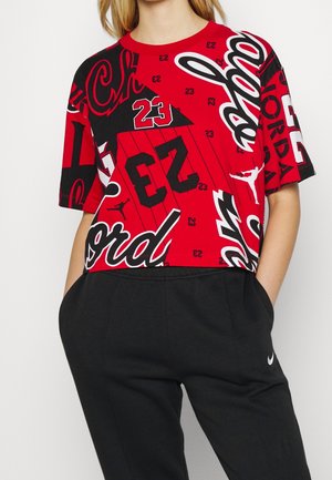 Personne portant un T-shirt court graphique Jordan rouge et noir et un pantalon de survêtement noir Nike, les mains dans les poches.