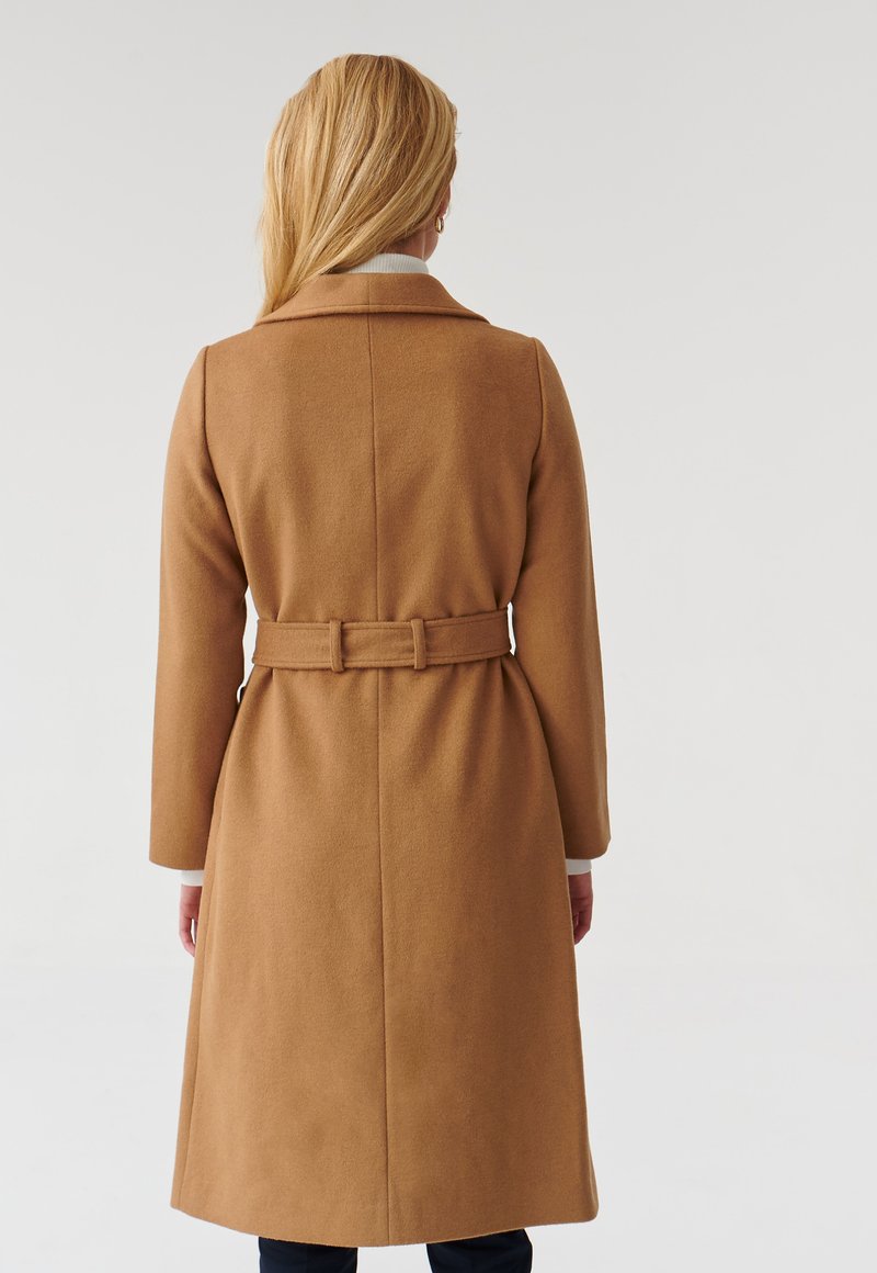 Cappotto di lana cammello con vita aderente, maniche lunghe e cintura posteriore. Texture liscia e colletto classico ne valorizzano il design strutturato.