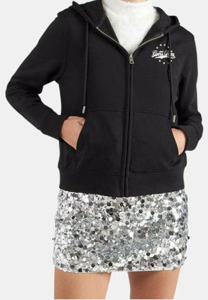 Czarna bluza z kapturem zapinana na zamek, z sznurkami, noszona na białym topie, w połączeniu z mini spódniczką ozdobioną srebrnymi cekinami, ręce włożone do kieszeni bluzy.