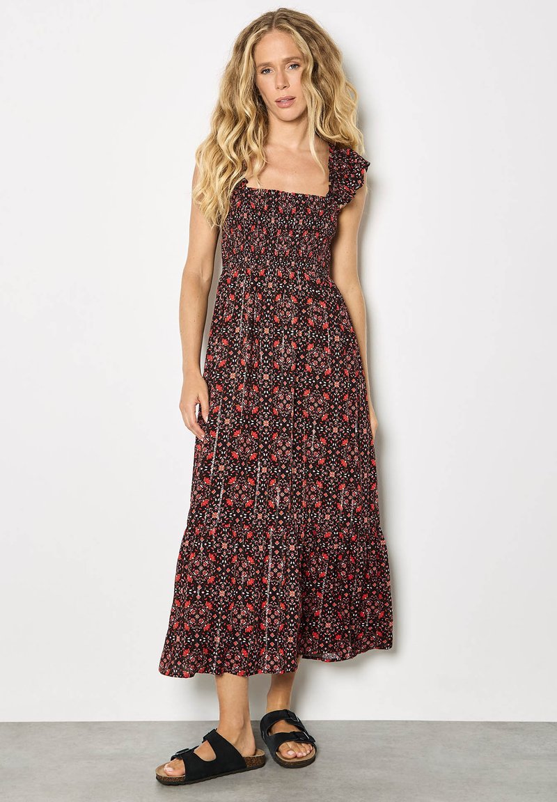 Femme aux longs cheveux blonds ondulés portant une robe midi noire sans manches à motifs floraux et des sandales noires à enfiler, debout contre un mur blanc.