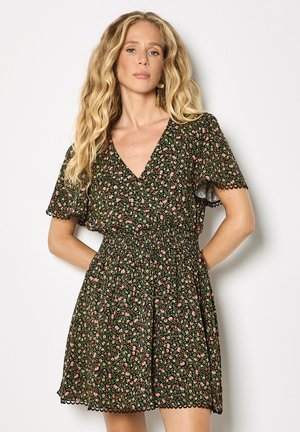 Kobieta z długimi blond włosami ubrana w krótką czarną sukienkę w kwiaty z rozkloszowanymi rękawami i falowanymi brzegami, stojąca na białym tle.