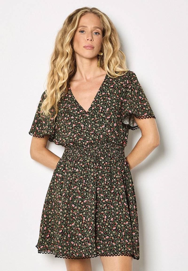 Femme aux longs cheveux blonds portant une robe courte noire à motifs floraux avec des manches flottantes et des bords festonnés, posant devant un fond blanc.