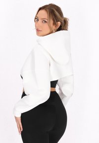 Korte witte hoodie met capuchon, brede mouwen en textuurstof, gecombineerd met zwarte high-waisted leggings.