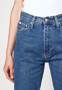 Höga blå jeans med klassisk passform, som har metallknappstängning, framfickor och blekt textur.