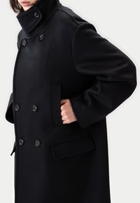 Svart yllekappa med hög krage, dubbelknäppt framsida med svarta knappar och sidofickor, med en slät och mjuk textur.