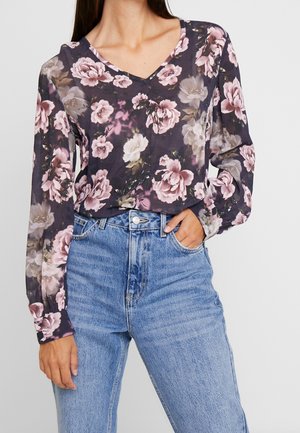 Kobieta w ciemnej, kwiecistej bluzce z długim rękawem, wpuszczonej w wysokowalne jasnoniebieskie dżinsy, z ręką w kieszeni.