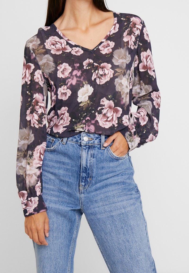 Femme portant un chemisier à manches longues à fleurs foncées, rentré dans un jean bleu clair taille haute, la main posée dans la poche.