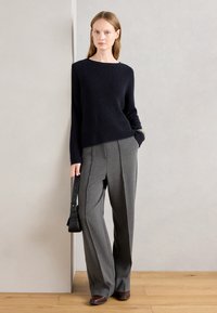 Maglione a costine navy con scollatura ampia, abbinato a pantaloni grigi a gamba larga. La modella tiene in mano una piccola borsa nera.
