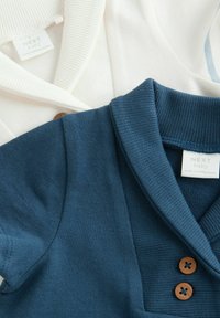 Twee baby polo shirts: één in crème met een geribde kraag en de andere in donkerblauw met een gestructureerde stof, voorzien van houten knoopdetails.