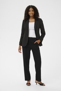 Žena s kudrnatými vlasy, oblečená v černém blejzru a kalhotách s bílým topem, pózující s jednou rukou v kapse a usmívající se.