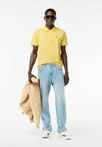 Camisa polo amarilla de manga corta, vaqueros de mezclilla azul claro, sosteniendo una chaqueta acolchada beige. Zapatillas blancas con detalles verdes. Lleva puestas gafas de sol.