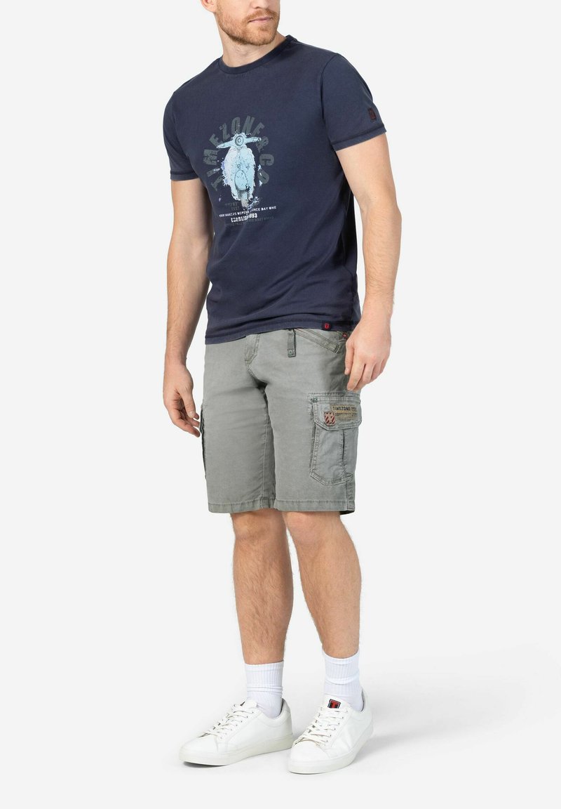Navy T-shirt with a graphic print, grey cargo shorts with side pockets, and white sneakers with textured fabric and laces.