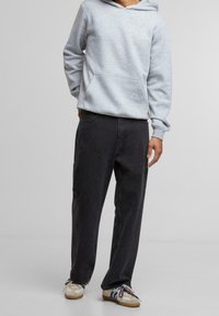 Persona che indossa un hoodie grigio chiaro, jeans neri casual e scarpe bianche con strisce blu, in piedi contro uno sfondo grigio uniforme.