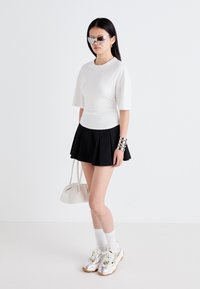Witte fitted top, zwarte geplooide rok, glanzende zilveren sneakers, witte sokken, zilveren zonnebril, zilveren manchettas, kleine witte handtas.