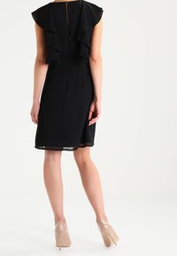 Czarna sukienka bez rękawów z falbanami na plecach, o długości do kolan, z prześwitującą warstwą na dole, zestawiona z cielistymi butami na wysokim obcasie.