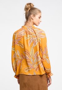 Blusa fluida a maniche lunghe color giallo senape con un motivo di foglie tropicali in rosa e crema. Presenta un collo arricciato e polsini elasticizzati.