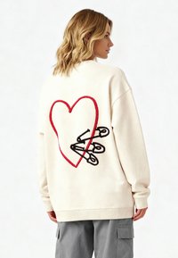 Felpa color crema con un grande cuore rosso e spille di sicurezza nere ricamate sul retro. Polsini e orlo a costine.