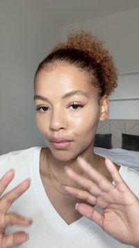 Osoba o kręconych, rudawo-brązowych włosach, ubrana w białą koszulę z dekoltem w serek, znajdująca się w neutralnym pokoju z łóżkiem i teksturowanymi poduszkami w tle.