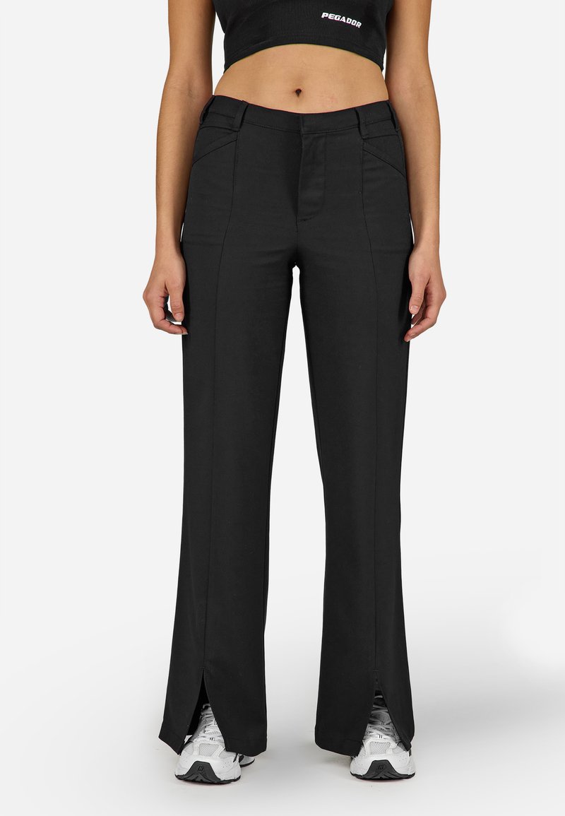 Žena v čiernych širokých nohaviciach s vysokým pásom, prednými švami a rozparkami na členkoch, kombinovaných s bielymi teniskami a skráteným čiernym topom.