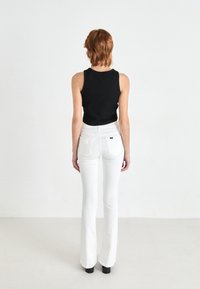 Jeans blancos de campana con bolsillos traseros y una camiseta sin mangas negra. La modelo está de pie con los brazos relajados a los lados, mostrando la vista trasera del conjunto.