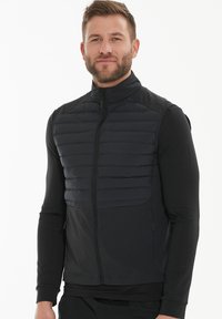 Schwarze Steppweste mit Reißverschluss auf der Vorderseite, ausgestattet mit gestepptem Textil im oberen Bereich und glattem Material im unteren Bereich. Stehkragen.
