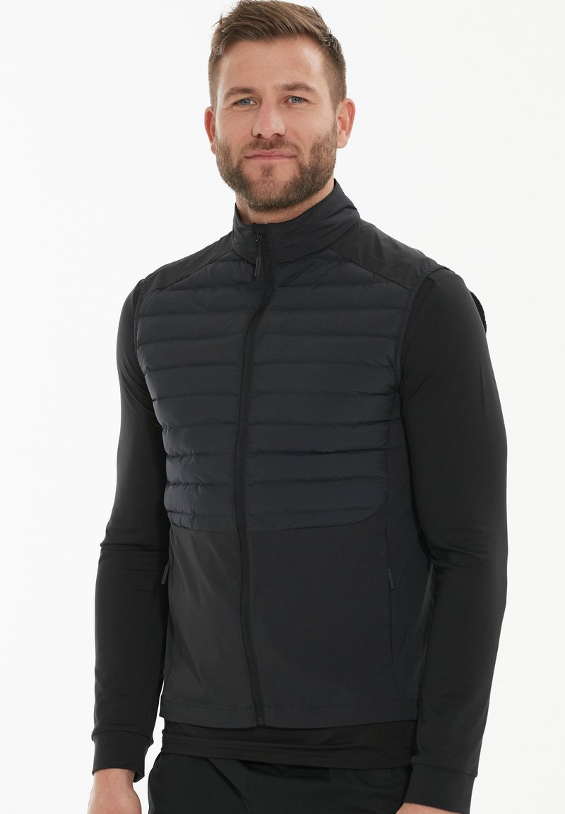 Schwarze Steppweste mit Reißverschluss auf der Vorderseite, ausgestattet mit gestepptem Textil im oberen Bereich und glattem Material im unteren Bereich. Stehkragen.