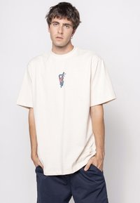 Mlad moški s kratkimi temnimi lasmi, oblečen v belo preveliko majico z majhnim modrim kačo in rožnato roko, ter temno modre hlače, z rokami v žepih.