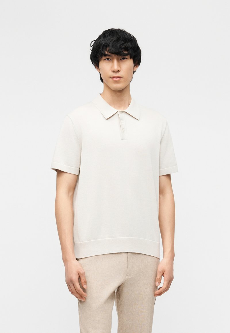 Jeune homme aux cheveux noirs bouclés portant un polo beige clair à manches courtes et un pantalon beige, debout devant un fond blanc uni.