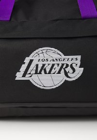 Zwarte stoffen tas met het witte logo van de Los Angeles Lakers en paarse handvatten die zichtbaar zijn aan de bovenranden.
