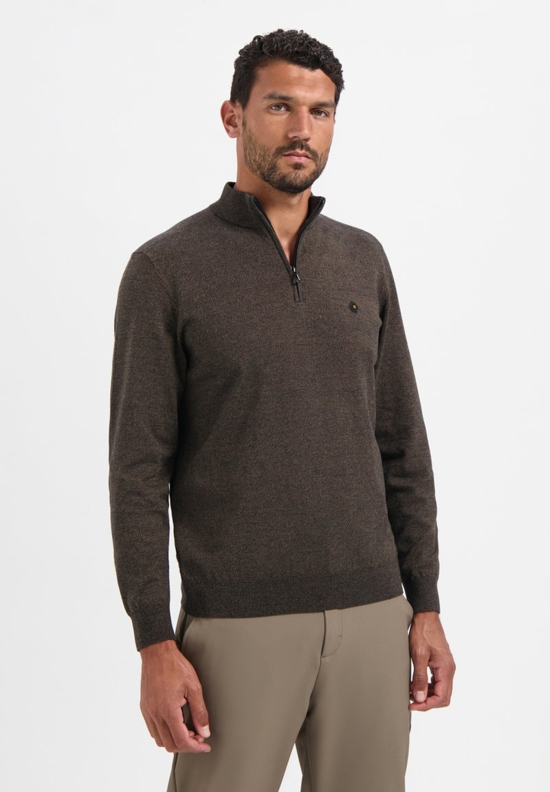 Bruine ritssluitingtrui van een gestructureerde stof, met een hoge kraag en een klein logo op de borst. Gestyled met beige broek.
