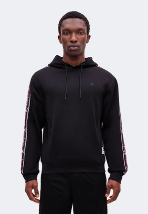 Jeune homme portant un sweat à capuche noir avec un ruban de marque rouge et blanc sur les manches, debout devant un fond gris clair uni.