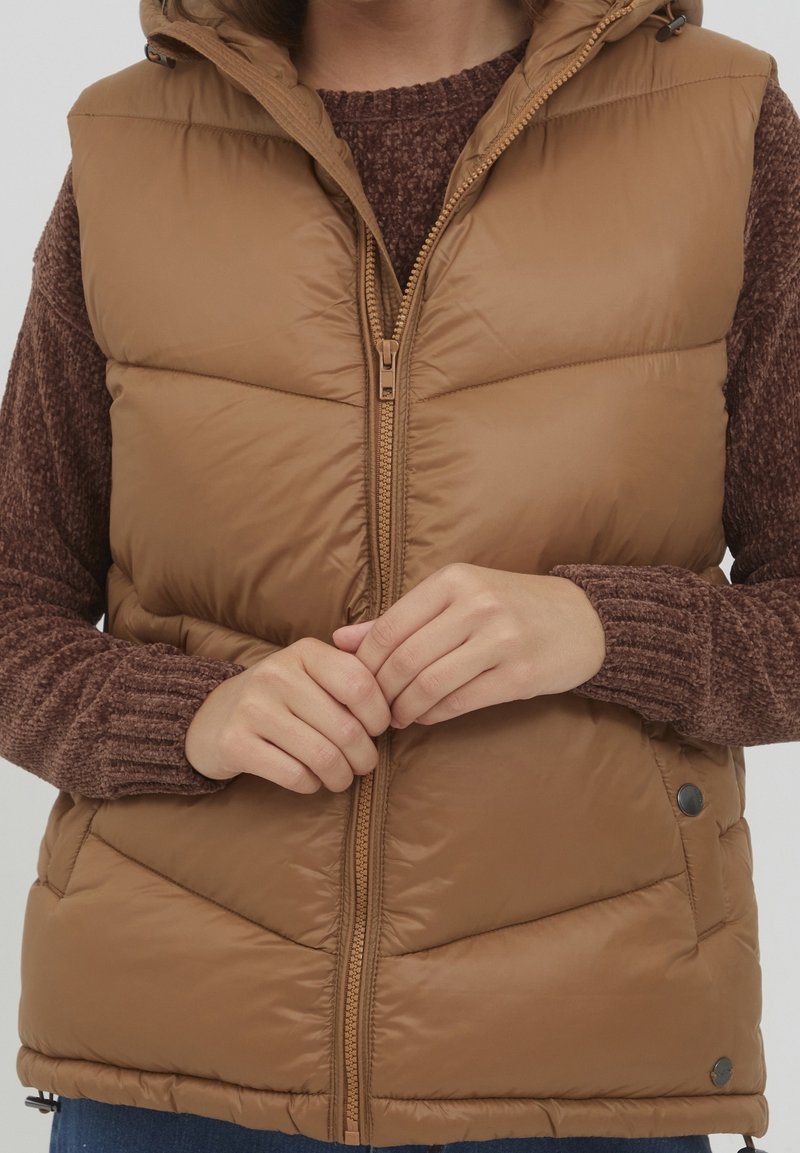 Personne portant une veste matelassée marron sur un pull marron texturé, mains jointes près de la fermeture éclair avant de la veste.