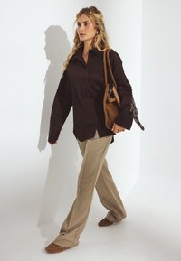 Sort skjorte med knapper, beige brede bukser, brune loafers og en brun skuldertaske, med en glat tekstur og en afslappet pasform.