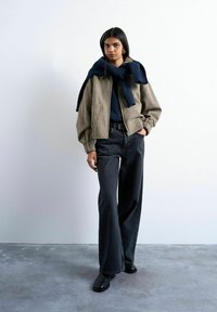 Femme debout portant une veste beige, un pull bleu marine drapé sur les épaules, un jean noir à jambes larges, des chaussures noires et des boucles d'oreilles créoles en or.