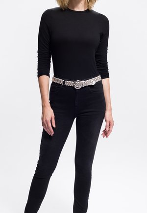 Donna che indossa una camicia a maniche lunghe nera e jeans neri a vita alta con una cintura intrecciata in argento, in piedi di fronte a uno sfondo bianco semplice.