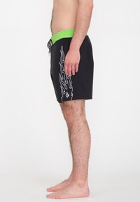 Bañador negro con cinturilla verde, que presenta un patrón de llamas blancas a lo largo del costado. Hecho de tela ligera, diseñado para la comodidad.