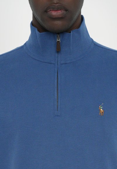 Blauwe ritstrui van gebreide stof met een hoge kraag. Bevat een klein logo van een ruiter op een paard. Rits met een leren lipje in bruin.