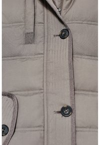 Lys grå polstret jakke med knaplukning foran, synlige stikninger og lynlås, med en blød tekstiloverflade og detaljeret tekstur.