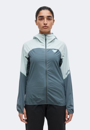Femme portant une veste à capuche zippée gris et bleu clair avec un logo sur la poitrine et la manche, debout face à nous avec les bras détendus.