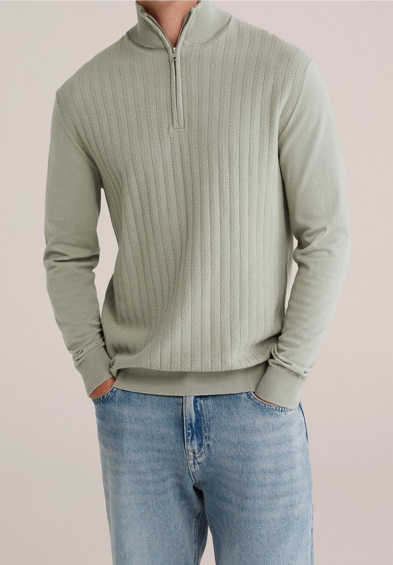 Homme portant un pull quart-zip texturé vert clair et un jean bleu clair, mains dans les poches, fond neutre.