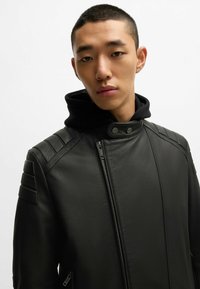 Giacca in pelle nera con un design strutturato, caratterizzata da accenti a coste sulle spalle, chiusura con zip e cappuccio staccabile nero.