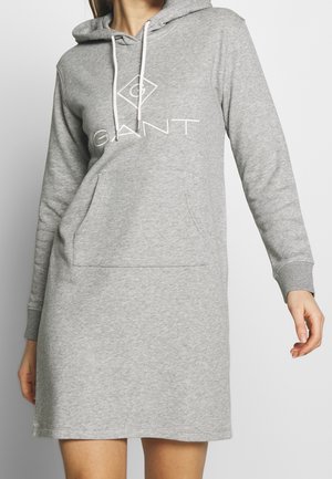 Vrouw die een lichtgrijze sweatdress met capuchon en een voorzak draagt, met het "GANT"-logo op de borst, staand tegen een effen achtergrond.
